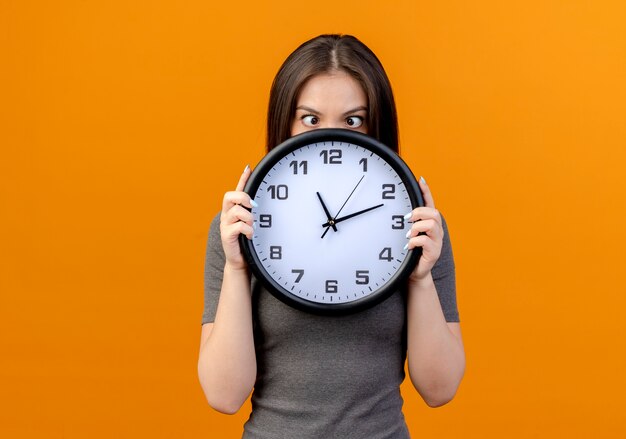 Beeindruckte junge hübsche Frau, die Uhr mit gekreuzten Augen hält und auf orange Hintergrund mit Kopienraum isoliert betrachtet