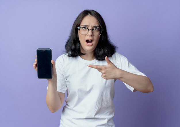 beeindruckte junge hübsche Frau mit Brille, die auf Handy zeigt und zeigt