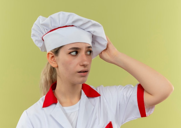 Kostenloses Foto beeindruckte junge hübsche köchin in kochuniform mit blick auf die seite, die hand auf ihren hut legt, isoliert auf grüner wand