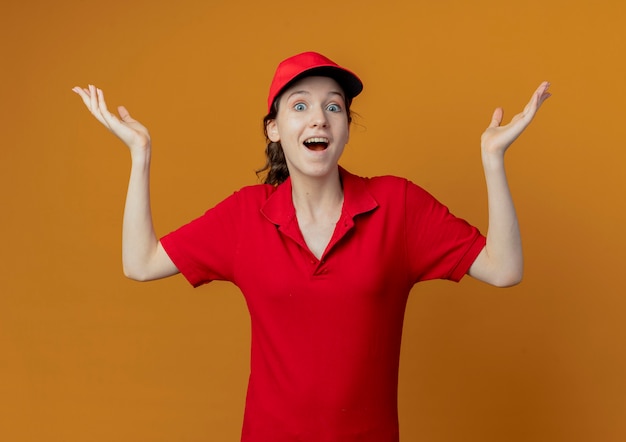 Beeindruckte junge hübsche Lieferfrau in roter Uniform und Kappe, die leere Hände lokalisiert auf orange Hintergrund zeigt