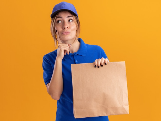 Beeindruckte junge hübsche Lieferfrau in Uniform legt Finger auf Gesicht und hält Papierpaket, das Seite betrachtet, die auf orange Wand lokalisiert ist