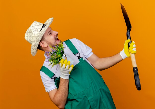 Beeindruckte junge hübsche slawische Gärtnerin in Uniform mit Hut und Gartenhandschuhen, die Spaten und Blumentopf halten und Spaten isoliert betrachten