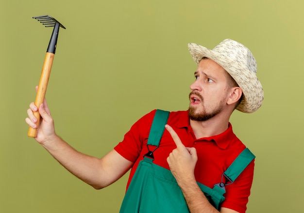 Beeindruckte junge hübsche slawische Gärtnerin in Uniform und Hut, die Rechen hält und auf sie lokalisiert auf olivgrüner Wand zeigt