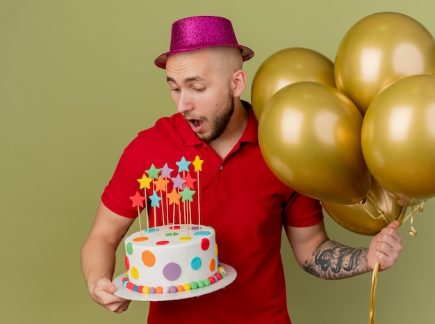 Beeindruckte junge hübsche slawische Partei Kerl, der Partyhut hält, der Ballons und Geburtstagstorte hält, die Kuchen lokalisiert auf olivgrünem Hintergrund betrachtet