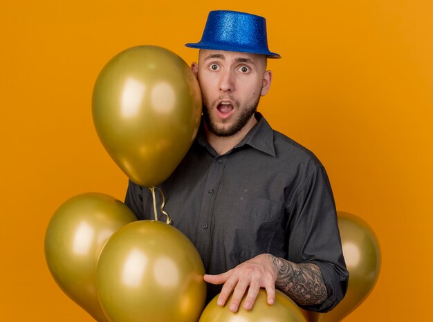 Kostenloses Foto beeindruckte junge hübsche slawische partei kerl, der partyhut trägt, der unter luftballons steht, die hand auf einen von ihnen setzen betrachten kamera lokalisiert auf orange hintergrund