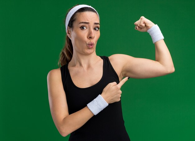 Beeindruckte junge hübsche sportliche Frau mit Stirnband und Armbändern, die nach vorne schaut und eine starke Geste macht, die auf ihre Muskeln zeigt, die auf grüner Wand isoliert sind?