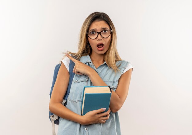 Beeindruckte junge hübsche Studentin, die Brille und Rückentasche hält Buch hält, das auf Seite lokalisiert auf Weiß zeigt