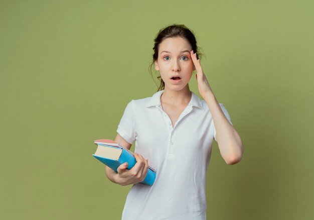 Beeindruckte junge hübsche Studentin, die Buch und Notizblock hält, die Hand auf Gesicht lokalisiert auf olivgrünem Hintergrund mit Kopienraum setzen