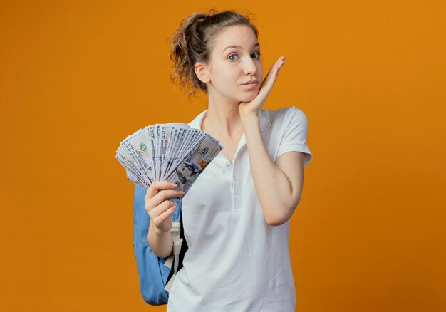 Beeindruckte junge hübsche Studentin, die Rückentasche hält, die Geld hält, das Kinn lokalisiert auf orange Hintergrund mit Kopienraum hält