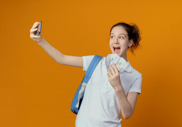 Beeindruckte junge hübsche Studentin, die Rückentasche hält, die Geld hält und selfie lokalisiert auf orange Hintergrund nimmt