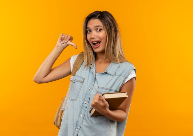 Beeindruckte junge hübsche Studentin, die Rückentasche hält, die offenes Buch hält, das auf sich zeigt, isoliert auf Orange