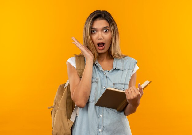 Beeindruckte junge hübsche Studentin, die Rückentasche hält, die offenes Buch zeigt, das leere Hand lokalisiert auf Orange zeigt