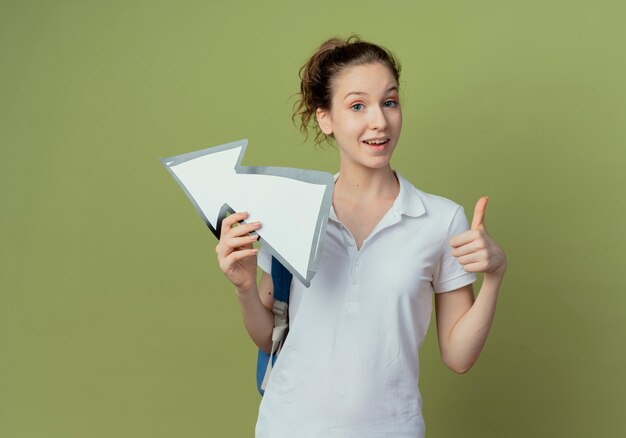 Beeindruckte junge hübsche Studentin, die Rückentasche hält, die Pfeilmarkierung hält, die zur Seite zeigt und Daumen oben lokalisiert auf olivgrünem Hintergrund mit Kopienraum zeigt