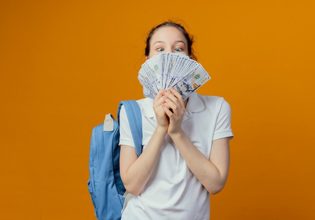 Beeindruckte junge hübsche Studentin, die Rückentasche hält und Geld lokalisiert auf orange Hintergrund mit Kopienraum trägt