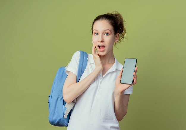 Beeindruckte junge hübsche Studentin, die Rückentasche trägt, die Handy und berührendes Gesicht lokalisiert auf grünem Hintergrund mit Kopienraum zeigt