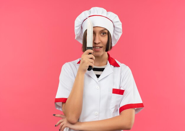 Beeindruckte junge Köchin in Kochuniform, die von hinten Messer schaut und Hand unter Ellbogen lokalisiert auf Rosa mit Kopienraum setzt