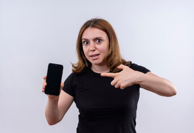 Beeindruckte junge lässige Frau, die Handy hält und auf isolierten weißen Raum zeigt