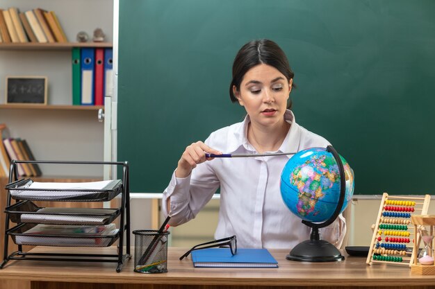 Beeindruckte junge Lehrerin, die am Tisch mit Schulwerkzeugen sitzt und den Zeiger im Klassenzimmer auf den Globus hält und aufsetzt