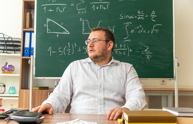 Beeindruckte junge Lehrerin mit Brille, die am Schreibtisch mit Schulmaterial im Klassenzimmer sitzt und die Hände auf dem Schreibtisch hält und zur Seite schaut