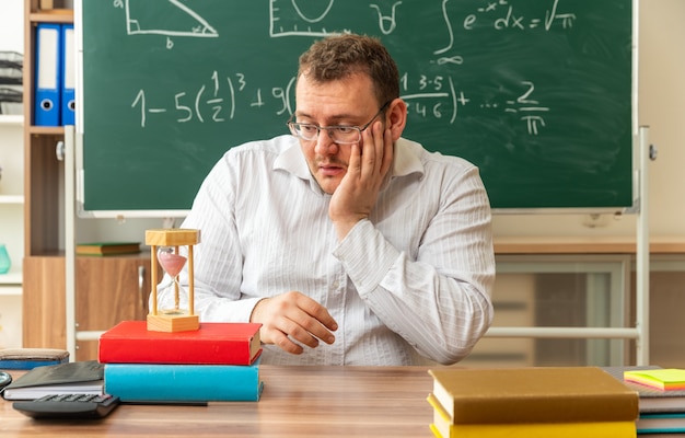 Beeindruckte junge Lehrerin mit Brille, die am Schreibtisch mit Schulmaterial im Klassenzimmer sitzt und die Hand auf dem Gesicht hält und auf die Sanduhr schaut