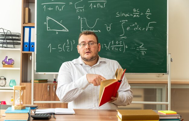 Beeindruckte junge Lehrerin mit Brille, die am Schreibtisch mit Schulmaterial im Klassenzimmer sitzt und ein offenes Buch hält, das auf die Vorderseite zeigt