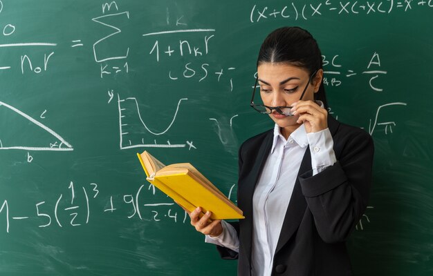 Beeindruckte junge Lehrerin mit Brille, die im Klassenzimmer vor der Tafel steht und ein Buch liest