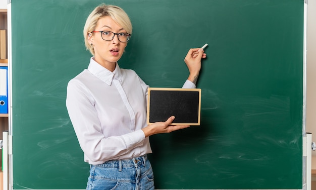 Beeindruckte junge Lehrerin mit Brille im Klassenzimmer, die in der Profilansicht vor der Tafel steht
