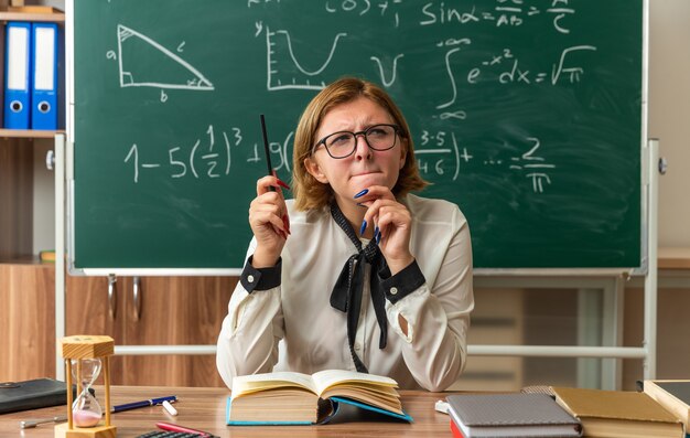 Beeindruckte junge Lehrerin mit Brille sitzt am Tisch mit Schulwerkzeugen, die im Klassenzimmer mit Bleistift gegriffenes Kinn halten