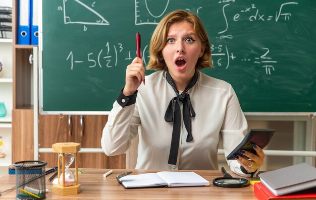 Beeindruckte junge Lehrerin sitzt am Tisch mit Schulmaterial, das einen Stift mit Taschenrechner im Klassenzimmer hält