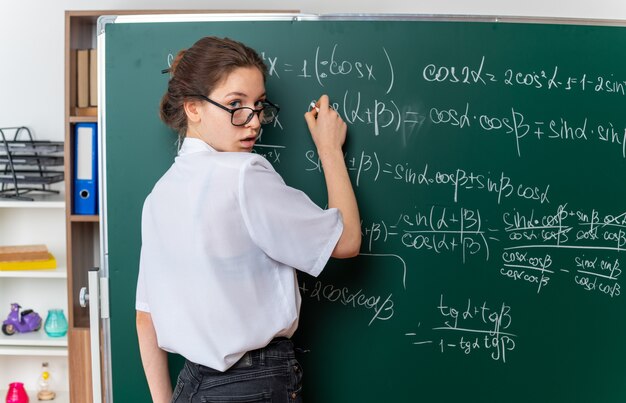 Beeindruckte junge Mathematiklehrerin mit Brille, die hinter der Tafel vor der Tafel steht und die Kreide hält, um mathematische Probleme im Klassenzimmer zu lösen?