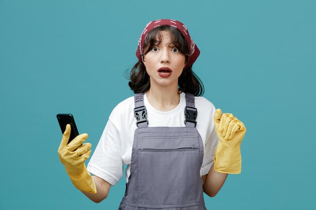 Beeindruckte junge Putzfrau mit einheitlichem Halstuch und Gummihandschuhen, die das Handy hält und die Hand in der Luft hält, die isoliert auf blauem Hintergrund in die Kamera blickt
