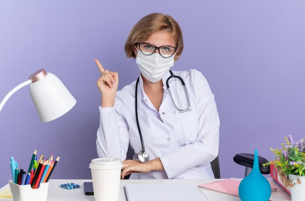 Beeindruckte junge Ärztin, die ein medizinisches Gewand mit Stethoskop und Brille mit medizinischer Maske trägt, sitzt am Tisch mit medizinischen Werkzeugen, die auf der blauen Wand isoliert sind