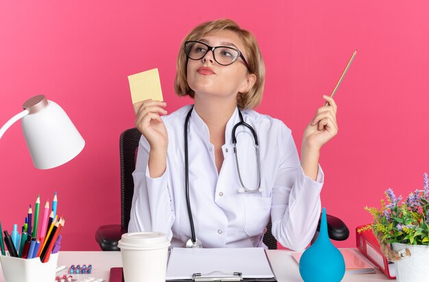 Beeindruckte junge Ärztin, die ein medizinisches Gewand mit Stethoskop und Brille trägt, sitzt am Schreibtisch mit medizinischen Werkzeugen, die Notizpapier mit Bleistift isoliert auf rosa Wand halten