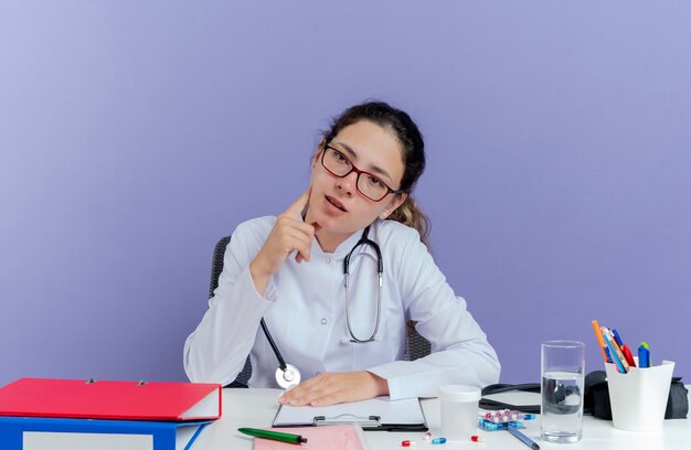 Beeindruckte junge Ärztin, die medizinische Robe und Stethoskop trägt, sitzt am Schreibtisch mit medizinischen Werkzeugen, die das Seitenberührungsgesicht lokalisieren