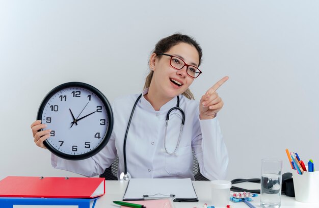 Beeindruckte junge Ärztin, die medizinische Robe und Stethoskop und Brille trägt, die am Schreibtisch mit medizinischen Werkzeugen sitzen, die Uhr halten und auf die Seite zeigen lokalisieren