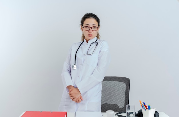 Beeindruckte junge Ärztin, die medizinische Robe und Stethoskop und Brille trägt, die hinter Schreibtisch mit medizinischen Werkzeugen stehen, die suchen, Hände zusammenzuhalten, isoliert