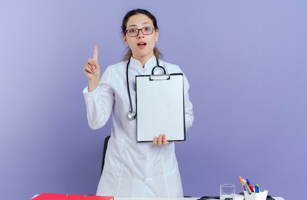 Beeindruckte junge Ärztin, die medizinische Robe und Stethoskop und Brille trägt, die hinter Schreibtisch mit medizinischen Werkzeugen stehen und Finger heben, der Klemmbrett lokalisiert zeigt