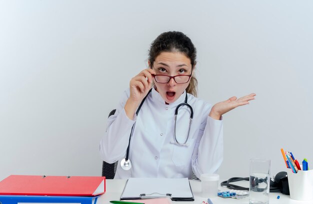 Beeindruckte junge Ärztin, die medizinische Robe und Stethoskop und Gläser trägt, die am Schreibtisch mit medizinischen Werkzeugen sitzen, die Gläser ergreifen, die leere Hand zeigen, die isoliert schaut