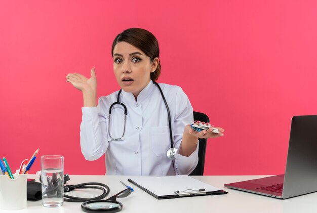 Beeindruckte junge Ärztin im medizinischen Gewand mit Stethoskop, das am Schreibtisch sitzt, arbeiten am Computer mit medizinischen Werkzeugen, die Pillen und Punkte mit der Hand zur Seite auf rosa Wand mit Kopienraum halten