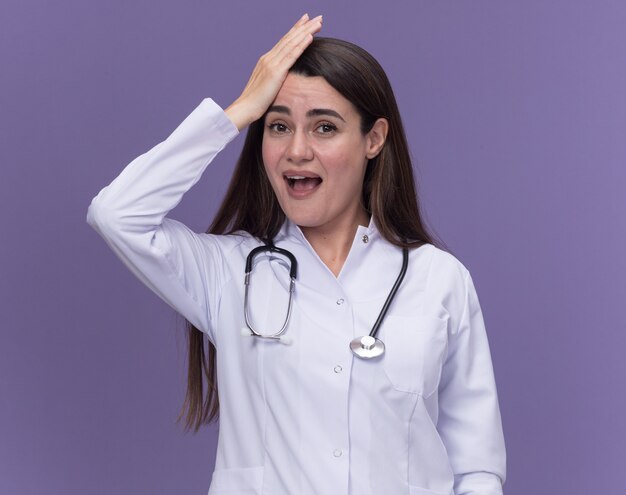 Beeindruckte junge Ärztin im medizinischen Gewand mit Stethoskop legt die Hand auf den Kopf und schaut auf die Kamera auf lila