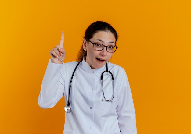 Beeindruckte junge Ärztin in medizinischer Robe und Stethoskop mit Brille zeigt nach oben isoliert