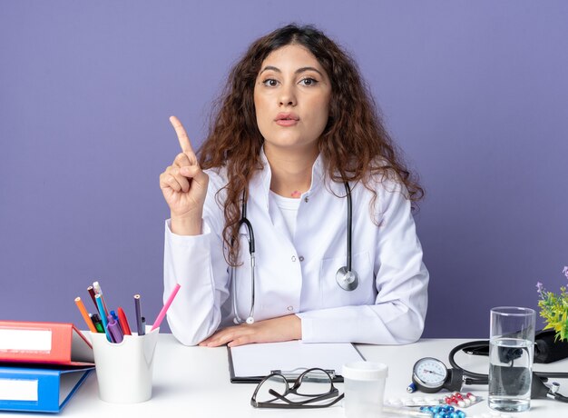 Beeindruckte junge Ärztin mit medizinischem Gewand und Stethoskop am Tisch sitzend mit medizinischen Instrumenten, die nach vorne schauen, isoliert auf lila Wand