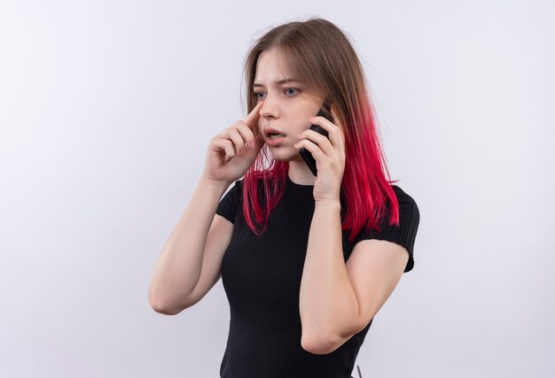 Beeindruckte junge schöne Frau, die schwarzes T-Shirt trägt, spricht am Telefon und legt ihre Hand auf Kinn auf isolierte weiße Wand