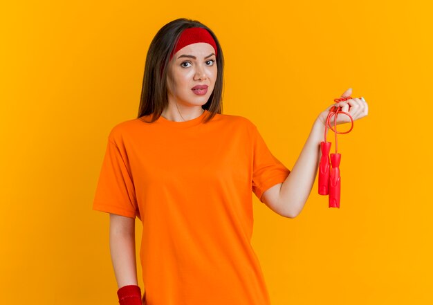 Beeindruckte junge sportliche Frau, die Stirnband und Armbänder hält, die Springseil suchen