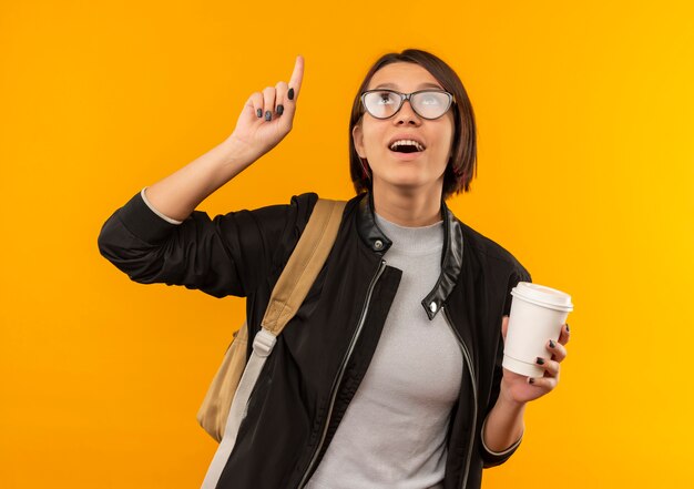 Beeindruckte junge Studentin, die Brille und Rückentasche hält, die Plastikkaffeetasse zeigt und lokalisiert auf Orange schaut