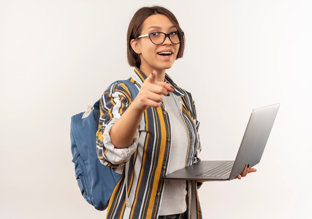 Beeindruckte junge Studentin, die Brille und Rückentasche hält Laptop hält, der auf lokal lokalisiert auf Weiß zeigt