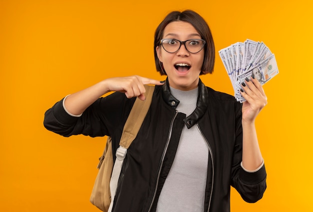 Beeindruckte junge Studentin, die Brille und Rückentasche hält und auf Geld zeigt, das auf Orange isoliert ist