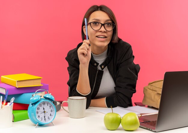 Beeindruckte junge Studentin, die eine Brille trägt, die am Schreibtisch hält Stift hält Hausaufgaben lokalisiert auf Rosa