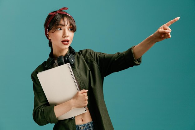 Beeindruckte junge Studentin mit Kopftuch und Kopfhörern um den Hals, die einen großen Notizblock mit Blick auf die Seite zeigt, die auf die Seite zeigt, die auf blauem Hintergrund isoliert ist