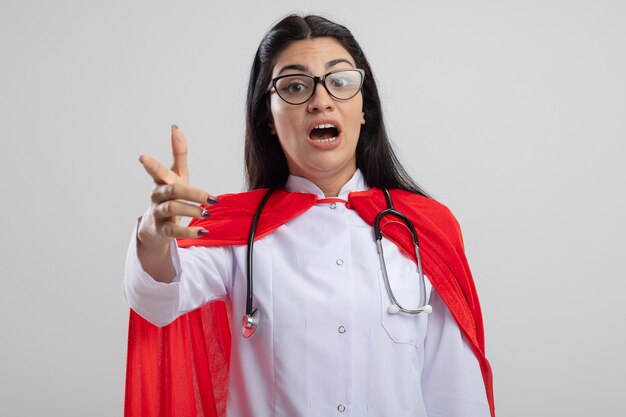 Beeindruckte junge Superfrau, die Brille und Stethoskop trägt und nach vorne zeigt, isoliert auf weißer Wand
